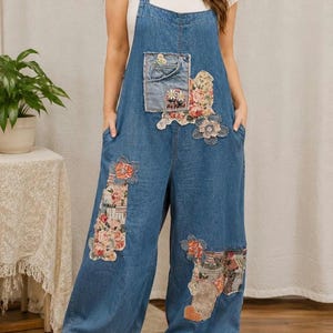 May include: Blue denim overalls featuring a wide-leg silhouette, a front chest pocket, and various floral and patterned fabric patches. The garment includes a white daisy trim along the bottom hems and a small patch with the text Be Kind.