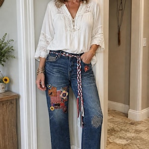 Puede incluir: Una blusa blanca de manga larga con detalles de encaje en el cuello y las mangas, combinada con unos vaqueros azul oscuro con varios parches de tela, un cinturón trenzado rojo, blanco y azul, y puños de tela estampada en el bajo.