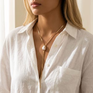 May include: A gold-toned lariat necklace featuring a flat, flexible snake chain and a central, irregular-shaped white pearl pendant. The necklace is worn around the neck, with the chain ends hanging down from the pearl setting in a Y-shape design.