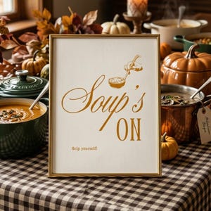 May include: A rectangular gold-framed sign with a white background featuring the gold text Soup's ON and the smaller text Help yourself. Above the text is a gold line drawing of a hand holding a ladle over a bowl of soup.