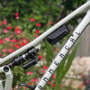 May include: A silver Commencal mountain bike frame featuring a black rear shock absorber and a small black rectangular storage bag mounted to the top tube. The frame displays the brand name Commencal in black lettering along the down tube.