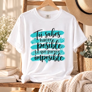 May include: A white short-sleeve t-shirt featuring a graphic design with horizontal turquoise brush strokes and black cursive text that reads: Tú sabes hacer posible lo que parece imposible, accented with small turquoise heart illustrations.