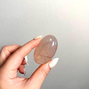 May include: A smooth, polished, oval-shaped rose quartz crystal with a translucent, pale pink hue. The stone features natural internal inclusions and a glossy, reflective surface that catches the light, showcasing its soft, muted color and rounded form.
