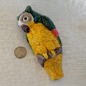 May include: A ceramic parrot figurine featuring a yellow chest, dark green wings, and a red tail accent. The bird is perched on a textured branch. A silver-colored coin is placed next to the figurine for scale, showing the relative size.