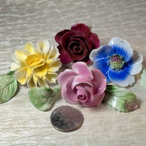 May include: A collection of four ceramic flowers including a yellow carnation, a deep red rose, a light pink rose, and a blue anemone, each with green leaves. A silver United States quarter coin is placed in the center for scale reference.
