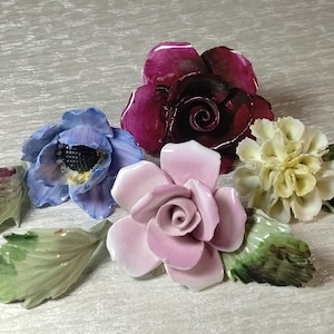 May include: A collection of ceramic floral ornaments featuring a deep burgundy rose, a light pink rose, a blue anemone with a dark center, and a cream-colored carnation, accompanied by several detached green ceramic leaves with detailed veining.