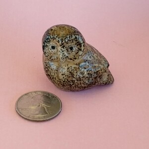 May include: A small ceramic owl figurine with a glossy, speckled brown and tan glaze. The owl has two dark, circular eyes and a rounded body. It is positioned next to a silver-colored United States quarter coin for scale.