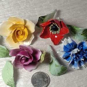 May include: A collection of five ceramic floral ornaments including a yellow rose, a purple rose, a red poppy, and a blue cornflower, accompanied by three detached green ceramic leaves and a silver-colored United States quarter coin for scale.