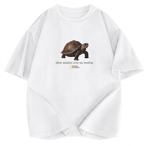 Puede incluir: Una camiseta blanca de manga corta con un gráfico de una tortuga marrón en el centro. Debajo de la tortuga, el texto dice "slow motion over no motion" seguido del logotipo de National Geographic en un marco rectangular amarillo.