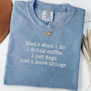 May include: A blue short-sleeved t-shirt with white text that reads: that's what i do i drink coffee i pet dogs and i know things. The shirt features a Comfort Colors brand label at the neckline and is paired with a gold-toned necklace with a small pendant.