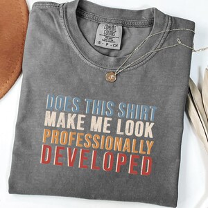May include: A charcoal gray short-sleeve t-shirt featuring the text "DOES THIS SHIRT MAKE ME LOOK PROFESSIONALLY DEVELOPED" in stacked lines of blue, cream, orange, and red. The shirt has a crew neckline and a Comfort Colors brand label.