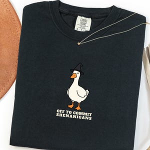May include: A black short-sleeved t-shirt featuring a graphic of a white duck wearing a black witch hat. Below the duck, the text reads OFF TO COMMIT SHENANIGANS in white capital letters. The shirt has a Comfort Colors brand label at the neckline.