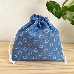 May include: A blue drawstring pouch featuring a repeating white circular starburst pattern. The bag is cinched at the top with thick white braided cotton cords that extend from the sides, creating a gathered fabric closure at the opening.