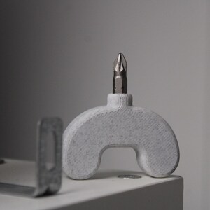 Peut inclure: Une poignée gris clair en forme d'arche avec une finition texturée, dotée d'un embout de tournevis métallique inséré au centre supérieur. À côté se trouve un support métallique plat en forme de L avec une découpe rectangulaire.