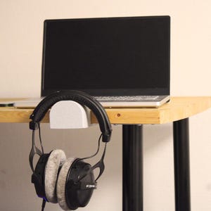 Op de afbeelding: Een witte plastic hoofdtelefoonhouder bevestigd aan de rand van een houten bureau. Een zwarte over-ear hoofdtelefoon met grijze velours oorkussens hangt aan de haak. Een zilveren laptop met een zwart scherm staat op het bureau achter de hoofdtelefoon.