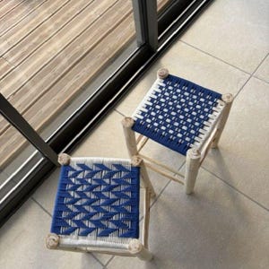 Peut inclure: Deux tabourets carrés avec des cadres en bois clair et des assises tressées. Une assise présente un motif à carreaux bleu et blanc, tandis que l'autre affiche un motif à chevrons bleu et blanc. Les tabourets ont quatre pieds et une structure simple.