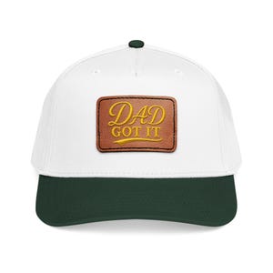 Puede incluir: Una gorra de béisbol blanca con visera verde oscuro y un botón verde oscuro en la parte superior. En el centro de la parte frontal hay un parche rectangular de cuero marrón con el texto amarillo bordado DAD GOT IT en letra cursiva.