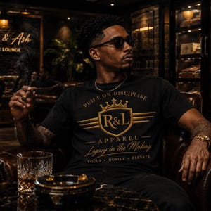 May include: A black t-shirt featuring a gold graphic print with a crown, a shield containing the letters R&L, and the text Built on Discipline, Apparel, Legacy in the Making, Focus, Hustle, Elevate. The design is centred on the chest area.