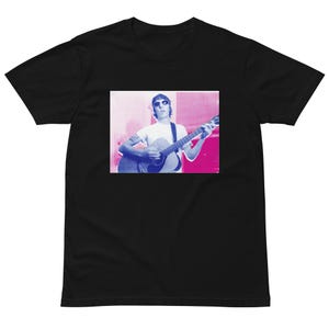 May include: A black short-sleeved t-shirt featuring a rectangular graphic print on the chest. The print displays a person wearing sunglasses and playing an acoustic guitar, rendered in a high-contrast color palette of bright pink and deep blue tones.
