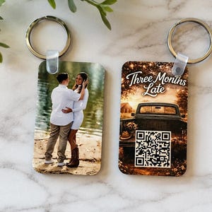 Puede incluir: Dos llaveros rectangulares con anillas de metal plateado. Uno muestra una fotografía de una pareja abrazándose al aire libre. El otro muestra una escena de atardecer con un camión, el texto Three Months Late y un código QR cuadrado.