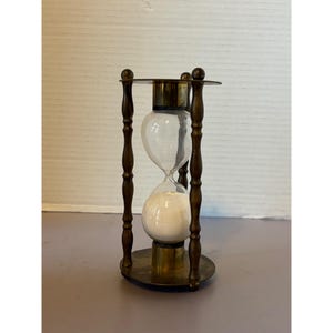 May include: An hourglass featuring a clear glass bulb containing white sand, held within a frame of four dark brass vertical pillars with decorative turned detailing, connected by circular brass top and bottom plates with a metallic finish.