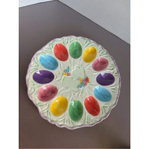 May include: A round ceramic egg serving platter featuring twelve oval indentations for eggs. The platter has a cream-colored base with a textured, light green leaf-like pattern and a scalloped edge. Each indentation is painted in vibrant colors.