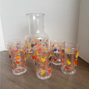 May include: A clear glass pitcher and four matching drinking glasses, each decorated with a red grid pattern and colorful illustrations of various fruits including grapes, cherries, lemons, pears, and watermelon slices on the exterior surface.
