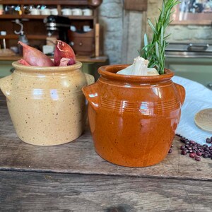 May include: Two ceramic pots with side handles, one in a speckled beige finish containing red shallots and one in a glossy terracotta orange finish containing a head of garlic and a sprig of fresh green rosemary, placed on a wooden surface.