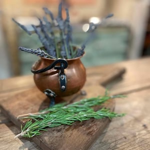 Peut inclure: Un petit chaudron en cuivre avec une anse en métal noir et trois pieds, contenant des brins de lavande séchée violette. Une planche à découper en bois se trouve sous le chaudron, avec des brins de romarin vert frais devant la planche.