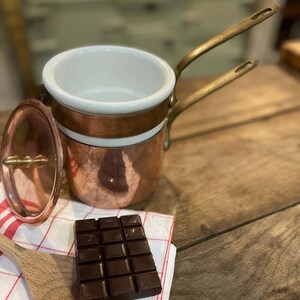 May include: A copper double boiler with a white ceramic insert and two brass handles. The set includes a matching copper lid with a brass finial. A dark chocolate bar and a wooden spoon are placed on a white cloth with red stripes.
