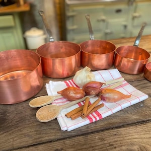 May include: A set of five graduated copper saucepans with metal handles arranged in a row. A white cloth with red stripes sits in front, holding a head of garlic, three shallots, two wooden spoons, and several cinnamon sticks.