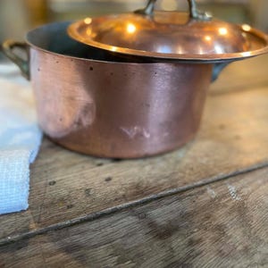 May include: A round copper pot with a matching lid and two side handles. The pot features a metallic copper exterior with a visible patina and a smooth, rounded shape. The lid has a central handle and rests partially atop the pot opening.