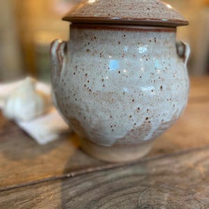 May include: A ceramic lidded jar with a rounded body and two side handles. The piece features a speckled off-white glaze with brown accents and a smooth, glossy finish. The lid has a small knob handle at the top, and the base is unglazed clay.