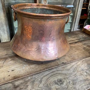 May include: A large, round, hammered copper pot with a wide, open top and a thick, rolled rim. The exterior surface features a textured, metallic finish with warm copper tones. Two small, circular metal handles are attached to the sides.