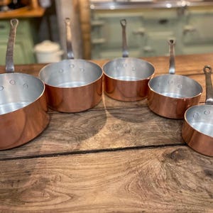 May include: A set of five graduated copper saucepans with silver-toned metal interiors and long, riveted metal handles. The pans are arranged in a row from largest to smallest, each featuring a polished copper exterior and a smooth, metallic lining.