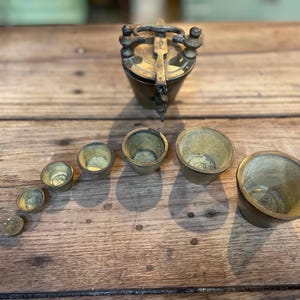 May include: A set of seven graduated brass nesting weights arranged in a curved line, accompanied by a lidded brass container with a locking handle mechanism. The metallic pieces feature a weathered patina and a conical shape for stacking.