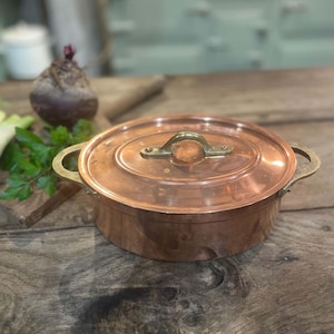 Op de afbeelding: Een ovale koperen pan met bijpassend deksel en twee koperen zijhandgrepen. Het deksel is voorzien van een centrale handgreep van messing die met klinknagels is bevestigd. De pan heeft een gepolijste metalen afwerking en een brede, ondiepe vorm.