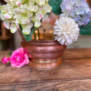 Könnte beinhalten: Eine gehämmerte Kupferschale mit einem messingfarbenen Rand und einem perforierten Metalleinsatz für Blumenarrangements. Die Schale enthält weiße und hellviolette Hortensienblüten sowie eine weiße Dahlie, mit einer leuchtend rosa Blume am Fuß.