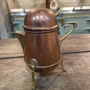 Op de afbeelding: Een koperen theepot met een gehamerde textuur, voorzien van een afgerond deksel, een gebogen tuit en een messing handvat. De theepot rust op drie slanke messing poten die vanaf de basis uitsteken en zorgen voor een driepoot-achtige stand.