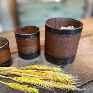 Könnte beinhalten: Drei zylindrische Holzbehälter in verschiedenen Größen, jeweils mit dunklen Metallbändern an den oberen und unteren Rändern. Das Holz hat eine natürliche braune Maserung und der größte Behälter enthält eine kleine Menge helles Material.