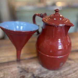 Pode incluir: Uma cafeteira de esmalte vermelho escuro com tampa a condizer e pega curva, posicionada ao lado de um funil vermelho escuro a condizer com interior azul claro. Ambos os itens apresentam um acabamento brilhante com desgaste visível.