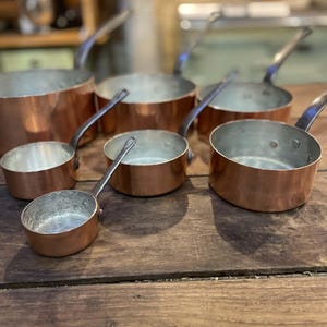 May include: A collection of six copper saucepans and pots of varying sizes, each featuring a polished copper exterior, a silver-toned metal interior lining, and a sturdy metal handle attached with rivets to the side of the round cooking vessel.