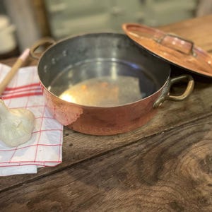 Könnte beinhalten: Ein runder Kupfertopf mit zwei seitlichen Messinggriffen und einem passenden Kupferdeckel. Das Innere besteht aus silberfarbenem Metall. Ein Holzlöffel, eine Knoblauchknolle und ein weißes Tuch mit roten Karos liegen neben dem Topf.
