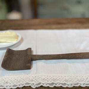 Op de afbeelding: Een vintage houten boterspaan met een rechthoekige kop met een diep gegroefd, geribbeld oppervlak voor het vormen en een lange, gladde, cilindrische steel. Het gereedschap is gemaakt van donker, verweerd hout met een zichtbare nerf.