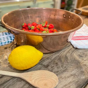 Puede incluir: Un cuenco grande de cobre con dos asas metálicas que contiene fresas rojas frescas. A su lado hay un limón amarillo brillante, un tarro de cristal con tapa de cuadros azules y blancos, una cuchara de madera y un paño blanco con rayas rojas.