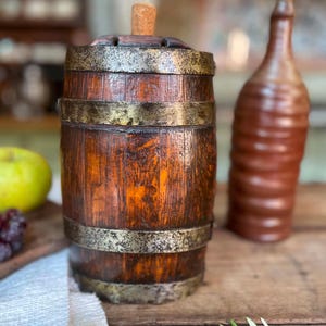 Könnte beinhalten: Ein zylindrisches Holzfass mit dunkelbrauner Oberfläche und verwitterten Metallbändern, die das Äußere umschließen. Die Oberseite ist mit einem zentralen Korkstopfen versehen. Das Fass hat eine vertikale Maserung und zeigt Gebrauchsspuren.