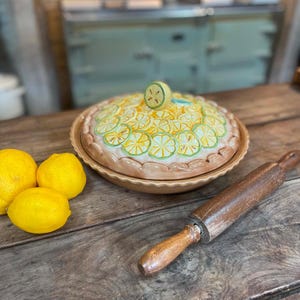 Peut inclure: Un plat à tarte en céramique avec un couvercle orné d'un motif de tranches de citron en relief et d'une poignée en forme de citron. Trois citrons jaunes entiers sont placés à gauche et un rouleau à pâtisserie en bois sombre est posé en diagonale.
