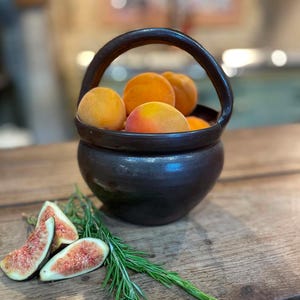 Op de afbeelding: Een donkerbruine keramische kom met een hoog gebogen handvat, gevuld met verschillende ronde oranje abrikozen. Naast de kom liggen drie gesneden verse vijgen met zichtbare roze binnenkanten en zaden, vergezeld door een takje groene rozemarijn.