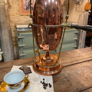 Op de afbeelding: Een gepolijste koperen koffiepercolator met een messing handvat en frame, voorzien van een centrale cilindrische behuizing, een scharnierend deksel en een zijhandgreep. Daarnaast staat een wit porseleinen theekopje met gouden rand op een bijpassende schotel, twee chocoladeblokjes en koffiebonen.