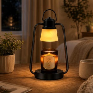 May include: A black metal candle warmer lamp with a handle and a frosted glass shade, featuring a glowing light directed downward onto a glass jar candle with a label that reads RE and HIBE, set on a circular base with a smooth matte finish.